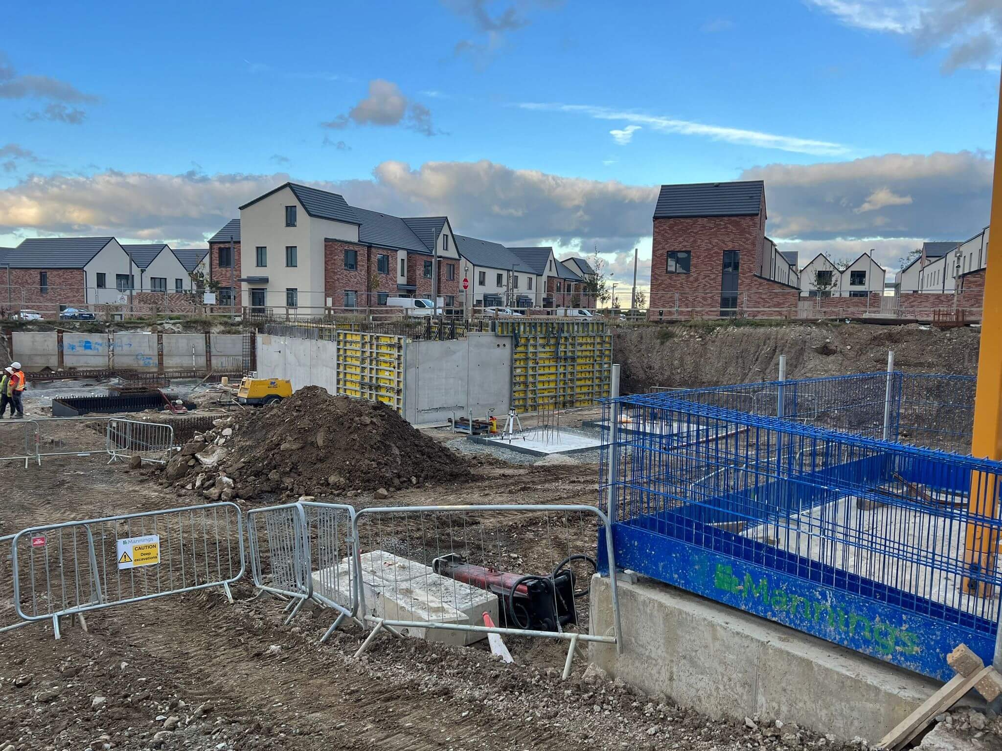 A construction site with an excavation pit, a fence, workers, and new houses in the background